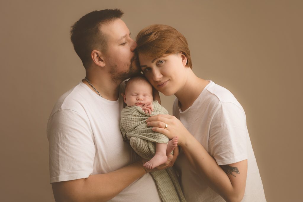 Ģimenes portrets ar tēti, mammu un viņu jaundzimušo meitiņu. Tētis tur meitiņu uz rokām un skūpsta māmiņu uz pieres.